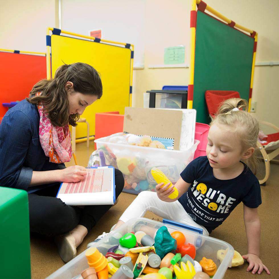 A student sits and records in a notebook while playing with a child